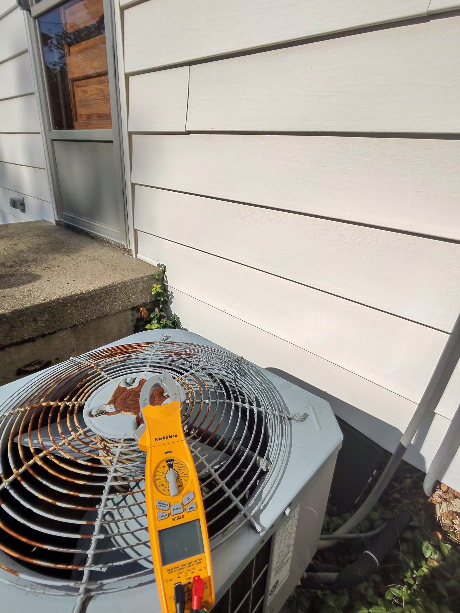 A yellow multimeter clipped to the fan of an outdoor air conditioning unit, with house siding in the background.
