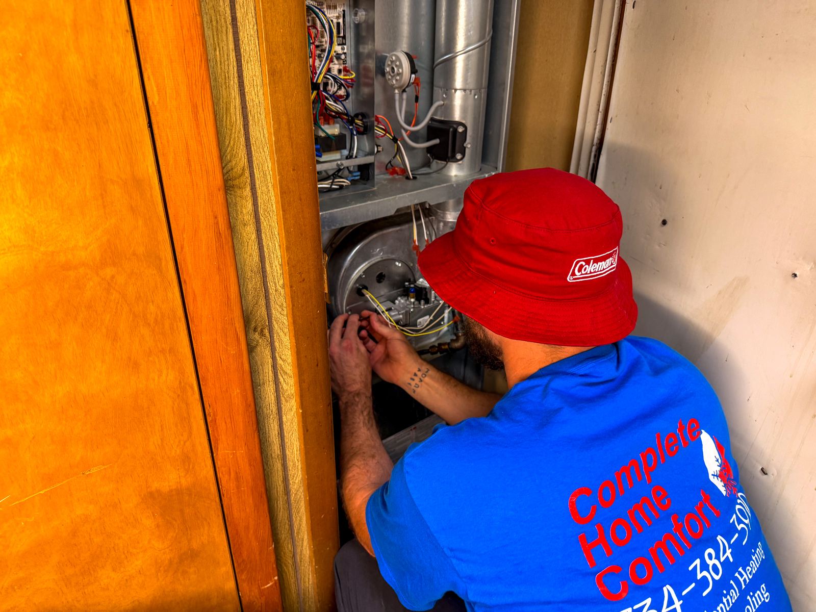 A technician in a red Coleman bucket hat and blue Complete Home Comfort shirt repairing the burner compartment inside an open furnace cabinet.