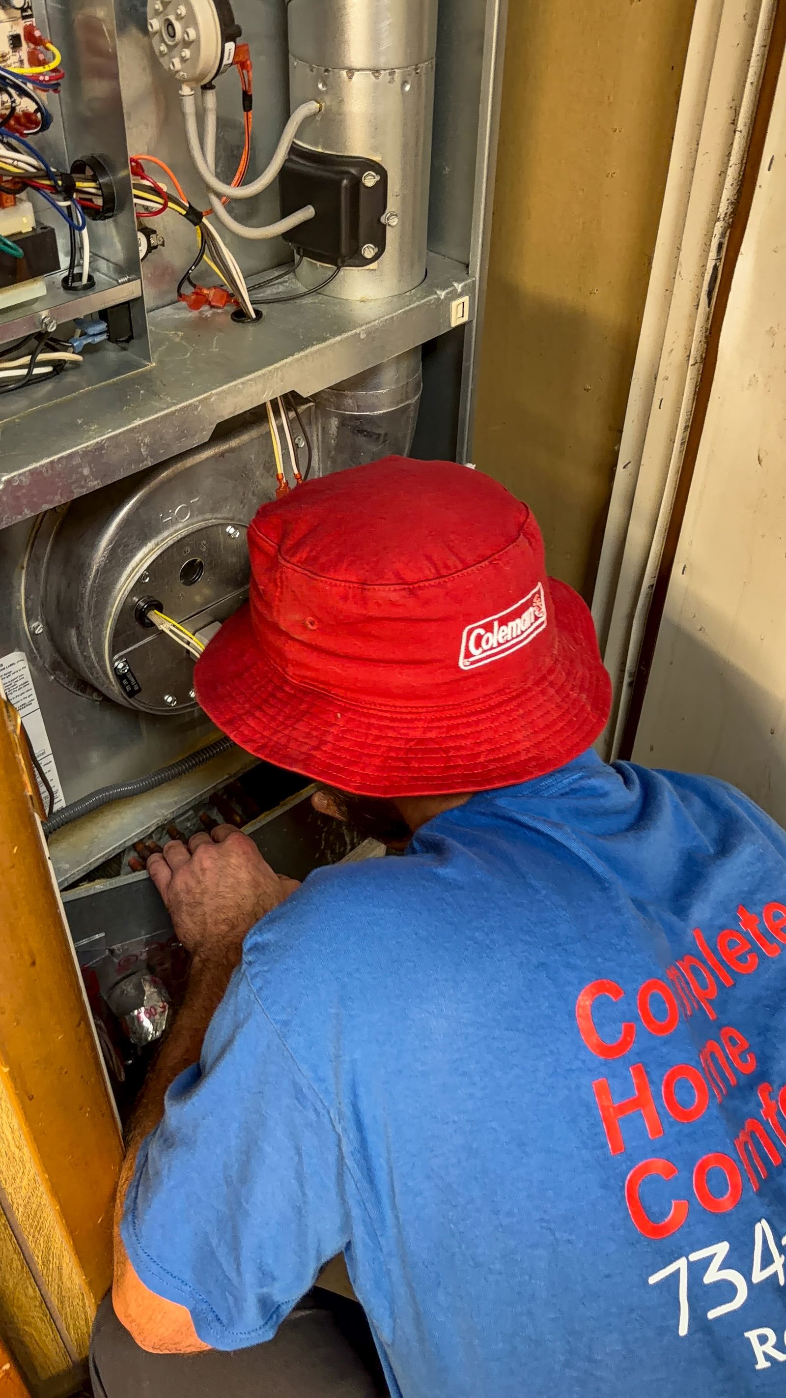 A technician in a red Coleman bucket hat and blue Complete Home Comfort shirt working inside an open furnace cabinet.