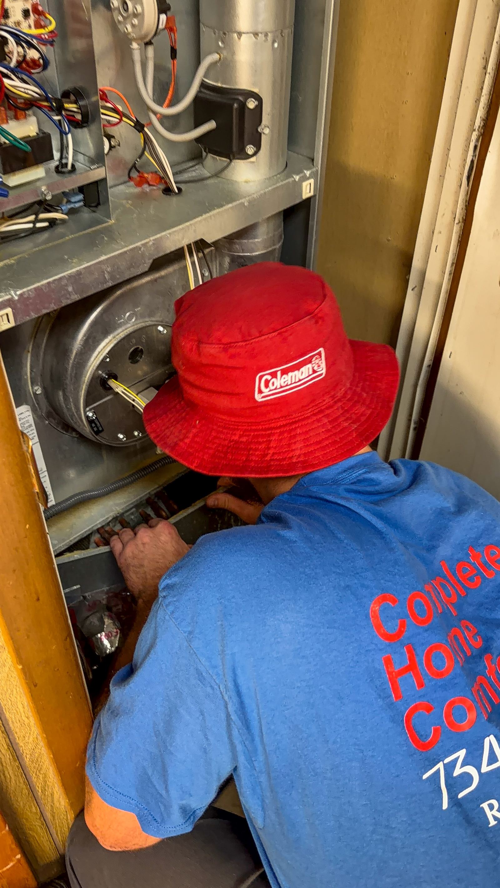 A technician in a red Coleman bucket hat and blue Complete Home Comfort shirt kneeling at an open furnace panel, seen from behind.