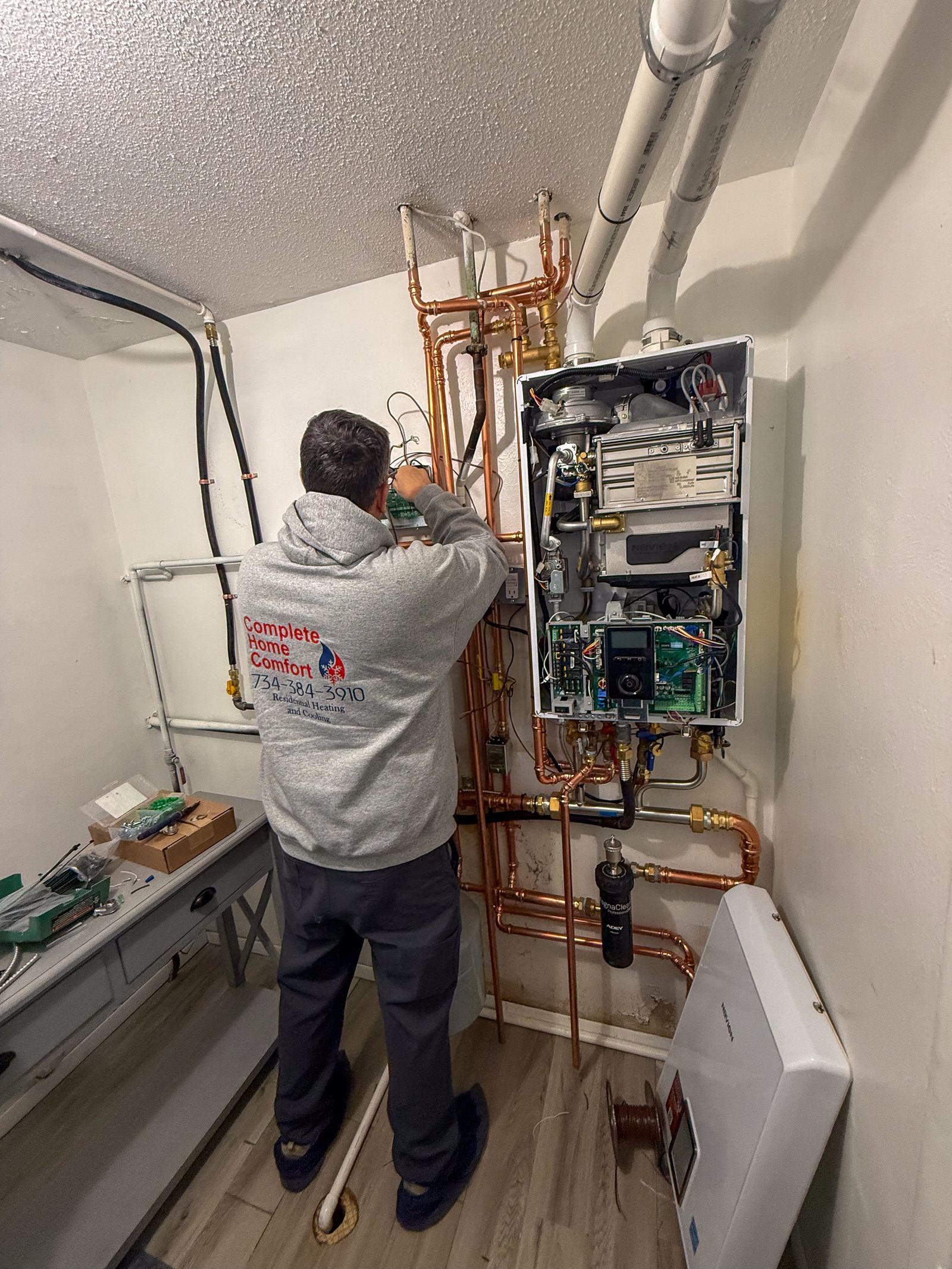 A technician in a Complete Home Comfort hoodie working on a wall-mounted tankless water heater with copper piping.